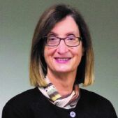 Linda Saris smiling at the camera, there is a gray background behind her