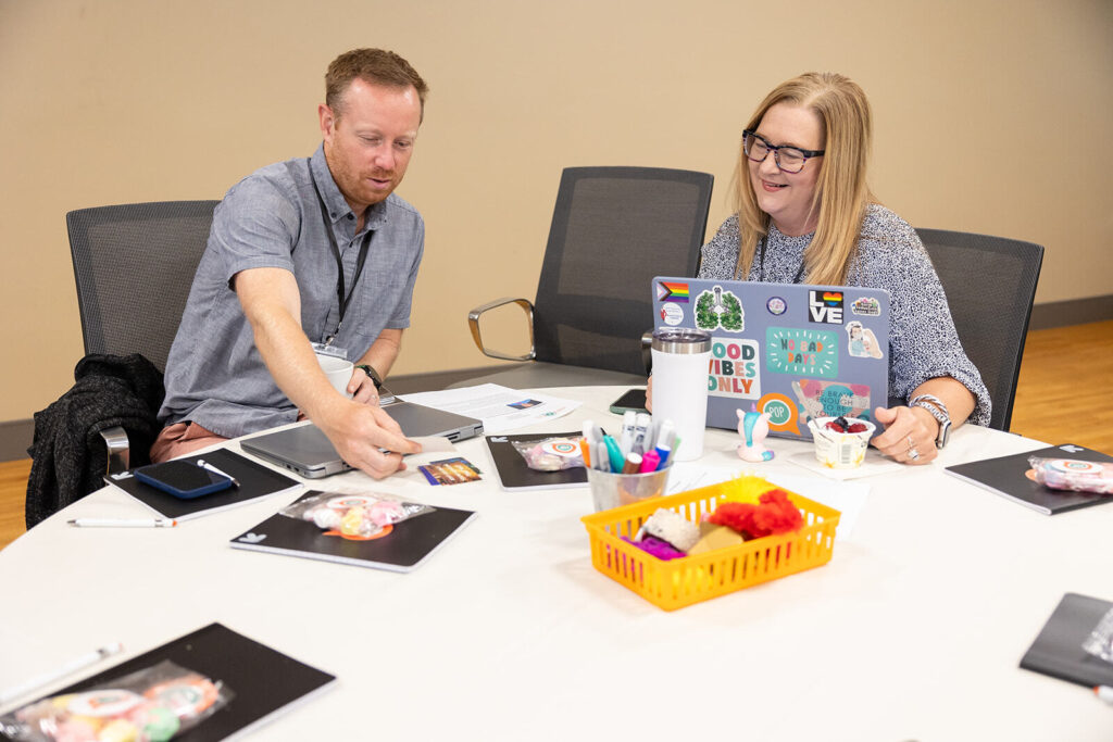 Two educators reviewing printed images around a table. One has their laptop open while the other reaches for a photo.