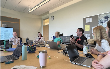 A group of educators is sitting around a conference table with their laptops open. One educator is speaking while the others listen.