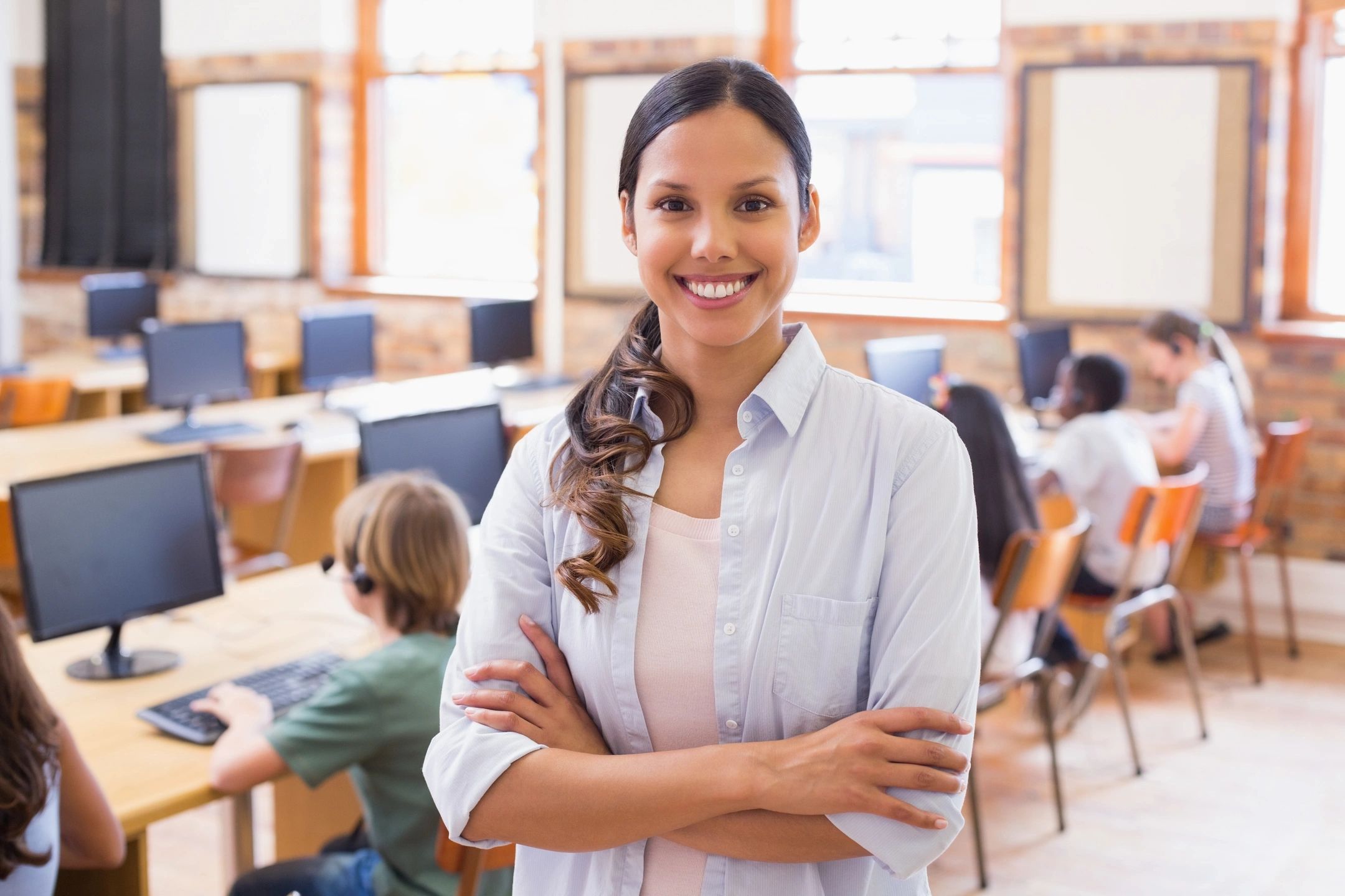 Teacher standing at the back of her classroom with her arms folded.