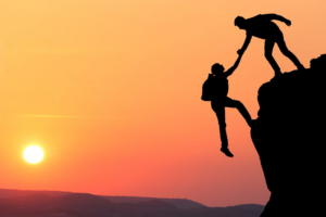 Person helping another person up a cliff in the sunset.